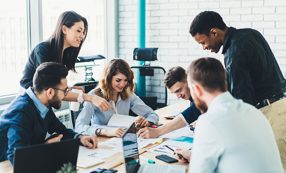 Successful group of male and female business analytics analyzing strategy for startup project during collaborating indoors, positive smiling colleagues enjoying cooperating near office desktop