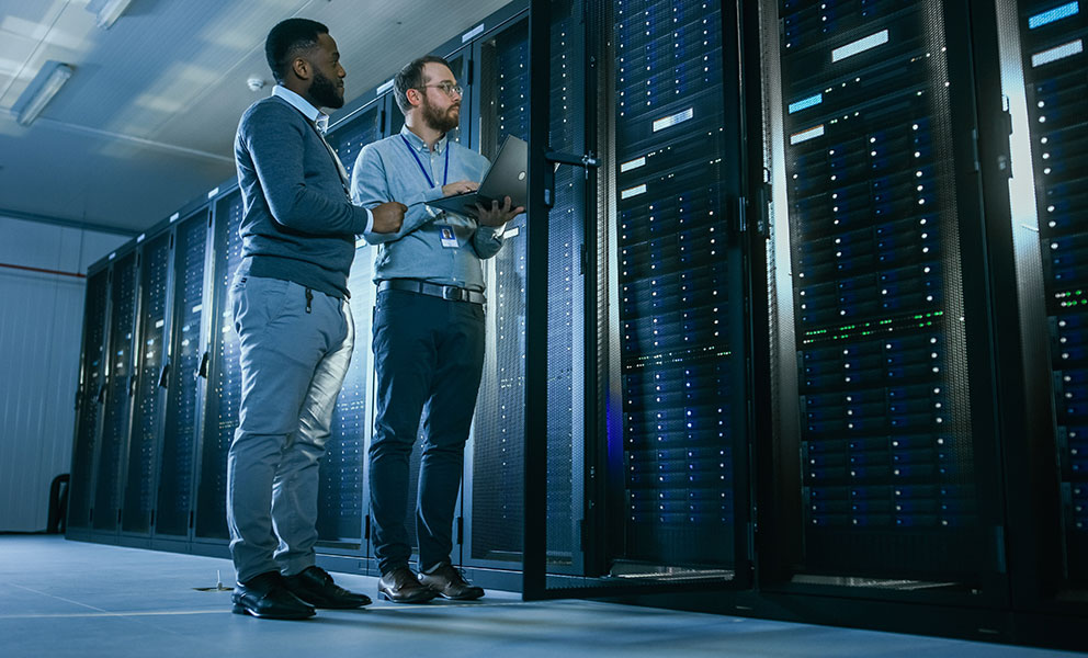 Bearded IT Technician in Glasses with a Laptop Computer and Black Male Engineer Colleague are Talking in Data Center while Working Next to Server Racks. Running Diagnostics or Doing Maintenance Work. 