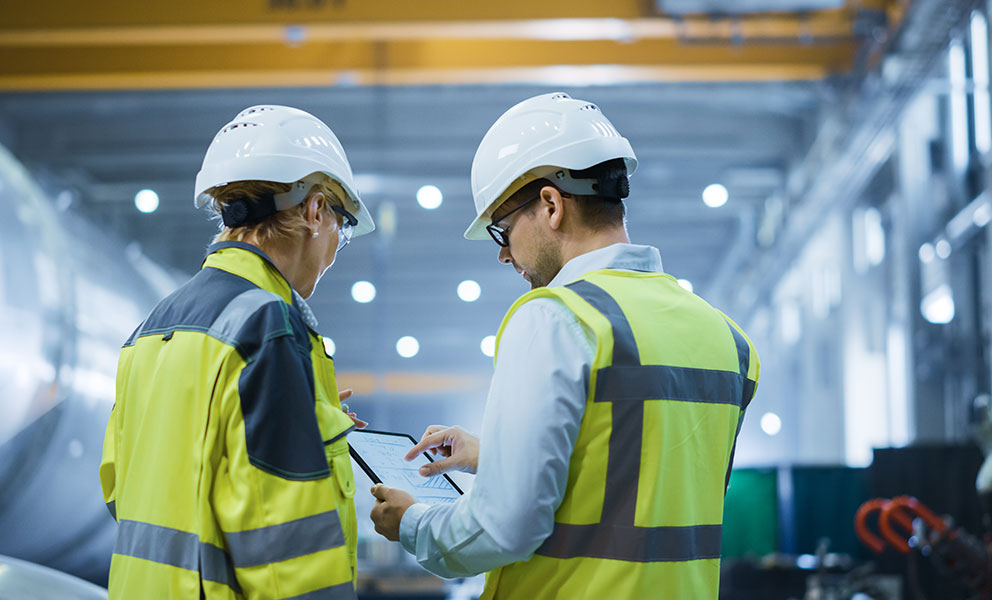 Two Heavy Industry Engineers Stand in Pipe Manufacturing Factory, Use Digital Tablet Computer, Have Discussion. Large Pipe Assembled. Design and Construction of Oil, Gas and Fuels Transport Pipeline